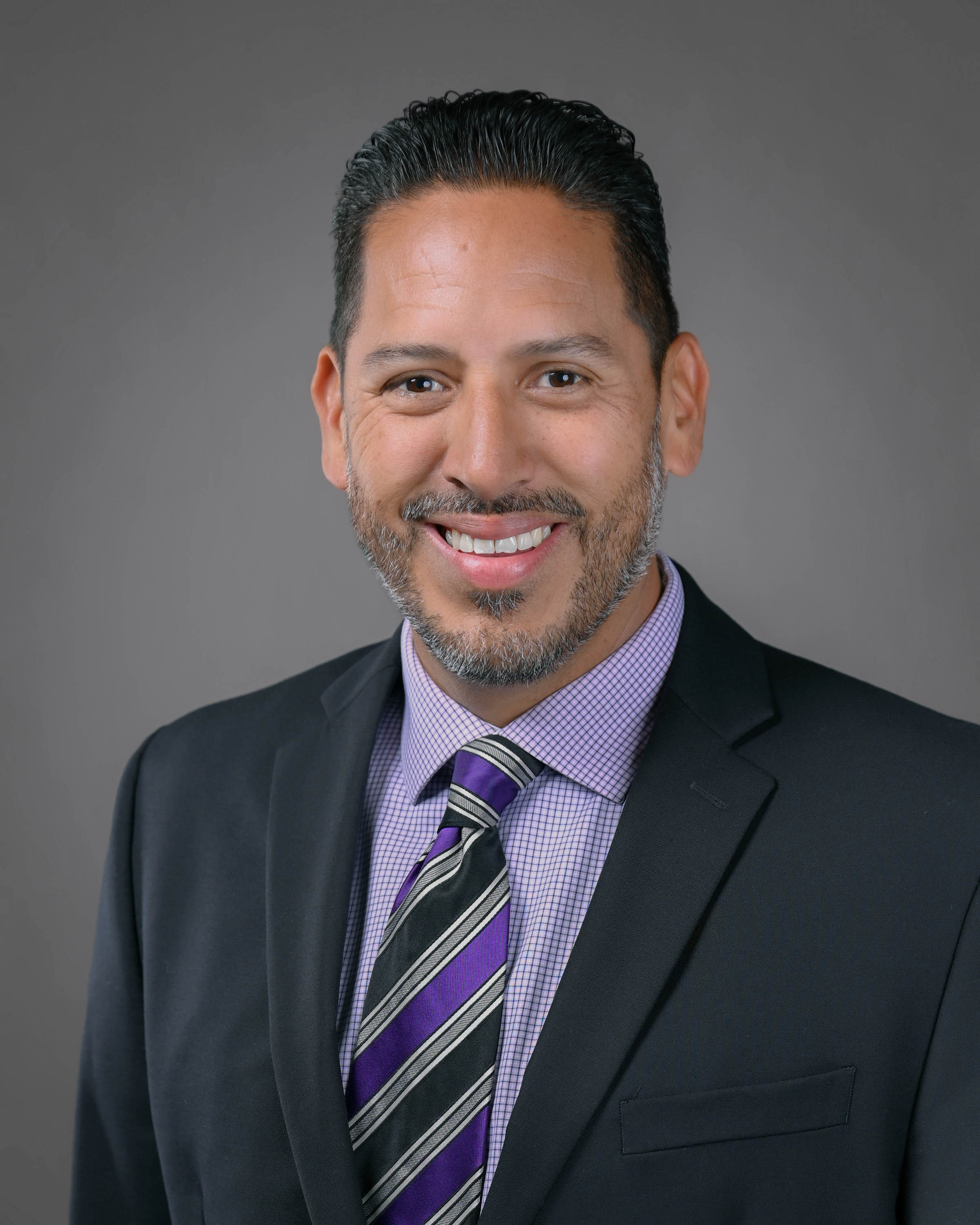 Professional headshot of a smiling man with short dark hair and a trimmed beard, wearing a black suit, purple checkered dress shirt, and striped purple tie against a gray studio background.
